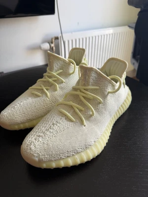 Yeezy Boost 350 V2 Butter sneakers - Snygga Yeezy Boost 350 V2 Butter sneakers från adidas. Skorna har en stickad ovandel i ljusgul Primeknit, matchande snören och en ribbad sula i samma ton. Ikonisk siluett med dragflik bak och bekväm Boost-dämpning.