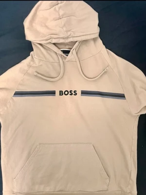 Boss tröja  - Snygg beige hoodie från BOSS med svart logotyp och två mörkgrå ränder över bröstet. Tröjan har huva med dragsko och en stor magficka framtill. Tillverkad i mjukt material som känns skönt mot huden. Perfekt för en avslappnad och trendig look.