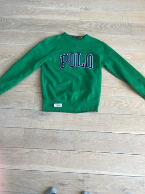 Grön sweatshirt från Polo Ralph Lauren - Snygg grön sweatshirt från Polo Ralph Lauren med stor POLO-text i vitt och blått på bröstet. Tröjan har rund halsringning, långa ärmar och ribbade muddar vid ärmslut och nederkant. Perfekt för en avslappnad och sportig stil. Hör er gärna av vid intresse.