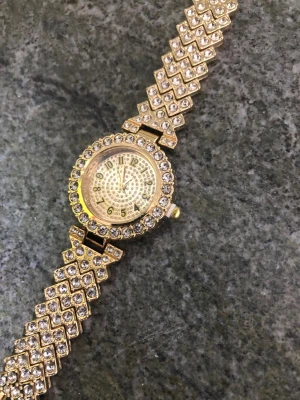 Klocka dam - Snygg guldfärgad klocka med glittrande stenar runt hela boetten och armbandet. Klockan har ett detaljerat mönster på urtavlan och siffror.Perfekt accessoar för dig som gillar bling och vill sticka ut.