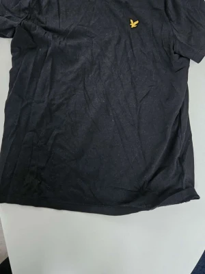 Svart t-shirt från Lyle & Scott - Klassisk svart t-shirt från Lyle & Scott med den ikoniska gula fågel-loggan broderad på bröstet. T-shirten har korta ärmar och är tillverkad i mjuk bomull för en bekväm känsla. Perfekt till jeans eller shorts.