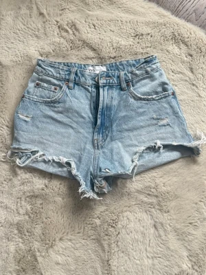 Ljusblå denimshorts med fransig kant - Säljer ett par ljusblå denimshorts från Zara med midrise midja och fransig, rå kant. Shortsen har klassisk femficksdesign, slitna detaljer och dragkedja framtill. Perfekta för sommaren och ger en avslappnad look. 
