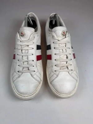 Vita Moncler sneakers med ränder - Snygga vita sneakers från Moncler i läder med klassiska röda och marinblå ränder på sidan. Skorna har lågt skaft, vita snören och Moncler-logga på plösen. Perfekta för dig som gillar stilrena och sportiga detaljer. Stl 44 äkta!