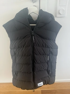 Svart dunväst med huva från Khujo - Svart Khujo-väst i storlek M. Knappt använd och i mycket fint skick. Perfekt för dig som vill ha en väst av hög kvalitet. Storlek M . Hör av dig vid frågor!  Pris: 750kr, kan diskuteras.