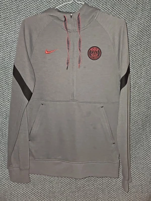 Grå PSG hoodie från Nike - Snygg grå hoodie från Nike med PSG-logga och röd swoosh på bröstet. Tröjan har huva med dragsko, halv dragkedja och en stor magficka. Svarta detaljer på ärmarna ger en sportig vibe. Perfekt för dig som gillar fotboll och streetwear. Är öppen för prisförslag vid snabb försäljning.