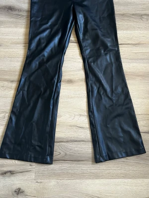 Svarta utsvängda fuskskinnbyxor - Snygga svarta byxor i fuskskinn med utsvängda ben. Byxorna har en hög midja och en glansig finish som ger en trendig look. 