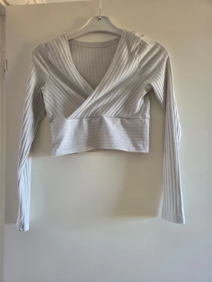 Vit omlottopp med ribbade detaljer - Snygg croppad omlottopp i vitt med ribbade detaljer och lång ärm. Toppen har en v-ringad omlottdesign framtill och är tillverkad i ett mjukt, stretchigt material som sitter bekvämt. Perfekt att styla med höga jeans eller kjol.