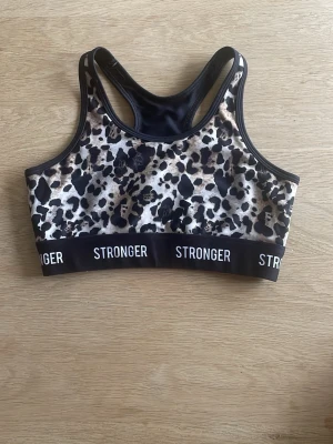 Leopardmönstrad sport-bh från Stronger - Sport-bh från Stronger med leopardmönster i svart, beige och brunt. Bred resår med logotypen STRONGER runt om. Racerback-modell med snygg cut-out i ryggen. Tillverkad i stretchigt funktionsmaterial som passar perfekt till träning.
