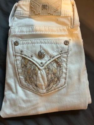 Vita Miss Me jeans med paljettdetaljer - Snygga vita jeans från Miss Me med dekorativa bakfickor prydda med silverfärgade paljetter och broderier. Jeansen har tydliga sömmar, silverknappar och ett emblem på linningen. Perfekta för dig som vill sticka ut med coola detaljer.
