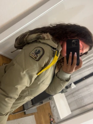  Parajumpers - Säljer en beige lång dunjacka från Parajumpers med ikonisk gul rem vid kragen och patch på ärmen. Jackan har en stor luva med fuskpäls och praktiska fickor. Perfekt för kalla vinterdagar och riktigt snygg streetstil. S/M
