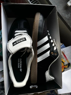 Adidas Wales Bonner svart  - Klassiska Adidas Samba OG sneakers i svart mocka med vita läderdetaljer och de ikoniska tre ränderna på sidan. Skorna har en brun gummisula och vita snören. Perfekt för dig som gillar retrostil och streetwear.
