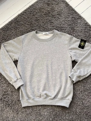 Grå sweatshirt från Stone Island - Snygg grå sweatshirt från Stone Island med klassisk rund hals och ribbade muddar. Tröjan har den ikoniska svarta patchen med gult och grönt Stone Island-märke på vänster ärm. Perfekt basic-plagg för en clean och stilren look.