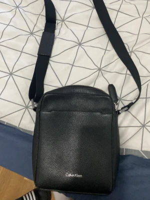 Svart axelväska från Calvin Klein - Snygg svart axelväska från Calvin Klein i strukturerat läder med justerbar axelrem och silverfärgad logga framtill. Väskan har dragkedja upptill och ett ytterfack för enkel åtkomst till småsaker. Perfekt för att bära det viktigaste.