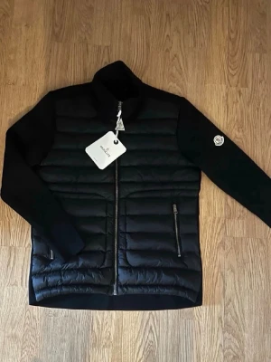 Svart dunväst från Moncler - Snygg svart dunväst från Moncler med quiltad front, två dragkedjefickor och hög krage. Västen har Moncler-logga på ärmen och stilren design. Perfekt för lager-på-lager och streetwear. Materialet är en mix av dun och syntet.
