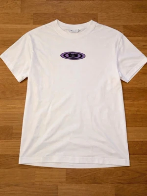 Vit t-shirt med lila tryck från Weekday - Säljer en vit t-shirt från Weekday med ett lila och svart grafiskt tryck på bröstet. Klassisk passform med rund hals och korta ärmar. Tillverkad i mjuk bomull som känns skön mot huden. Perfekt till jeans eller shorts.