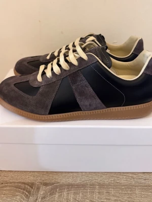Maison Margiela Replica sneakers grå - Snygga Maison Margiela Replica sneakers i grå mocka och svart läder med beigea skosnören och gummisula. Klassisk low-top siluett med retrovibbar och diskret logga på plösen. Perfekta för dig som gillar stilrena och tidlösa sneakers.