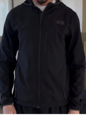  The North Face jacka  - Snygg svart vindjacka från The North Face med huva och dragkedja framtill. Jackan har två sidofickor med dragkedja och diskret logga på bröstet. Tillverkad i lätt och slitstarkt material, perfekt för blåsiga dagar.