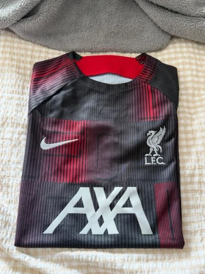 Liverpool FC träningströja Nike - Snygg Liverpool FC träningströja från Nike i svart med röda och vinröda detaljer. Tröjan har klubbmärke, Nike-logga och AXA sponsortryck på bröstet. Tillverkad i ett lätt och funktionellt material som passar perfekt till träning.