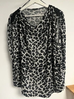 Leopardmönstrad topp med drapering - Säljer en snygg leopardmönstrad topp i svart och grått med draperad halsringning och rynkade axlar. Toppen har lång ärm och är gjord i ett mjukt, stretchigt material som känns skönt mot huden. Perfekt för dig som vill sticka ut lite extra.