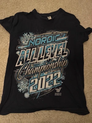 Svart t-shirt Nordic Championship 2022 - Cool svart t-shirt från SOL'S Imperial med stor tryckt text 'Nordic All Level Championship 2022' i vitt, blått och silver. Snygga snöflingor och grafiska detaljer framtill. Perfekt för dig som gillar sportiga statement-plagg.