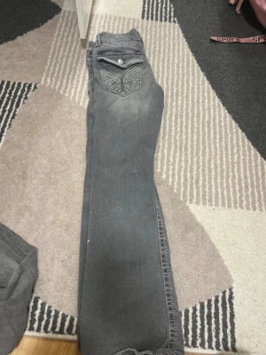 Grå jeans med broderad ficka - Säljer ett par grå jeans med snygg broderad bakficka och knappdetalj. Jeansen har rak passform och klassisk femficksdesign. Perfekta för dig som vill ha en stilren men ändå unik look. Betalning via vanlig swish! 