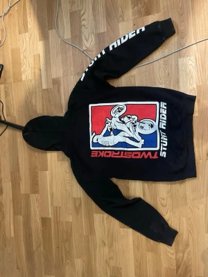 Svart Two Stroke hoodie med tryck - Svart hoodie från Two Stroke med stort tryck på ryggen av en stuntande motorcyklist i rött, vitt och blått. Framsidan har stor vit och röd logga samt ficka framtill. Texten 'STUNT RIDER' finns längs ena ärmen. Klassisk huva och mjukt material.