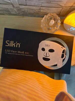 Silk'n LED Face Mask 100 - LED-ansiktsmask från Silk'n i vit plast med svarta detaljer. Masken har utskärningar för ögon, näsa och mun samt ett tillhörande kontrollverktyg. Används för anti-age och för att minska hudens ojämnheter. Perfekt för hemmabruk och enkel att använda.