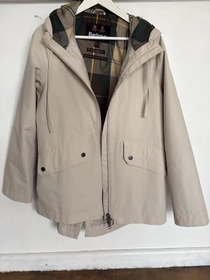 Beige regnjacka från Barbour - Stilren beige regnjacka från Barbour med huva och klassiskt rutigt innerfoder. Jackan har två stora fickor framtill med tryckknappar och dragsko i midjan för snygg passform. Perfekt för regniga dagar och enkel att matcha med olika outfits.