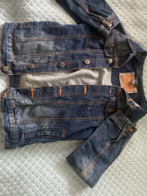 Klassisk blå jeansjacka - Säljer en klassisk blå jeansjacka med orangea sömmar och silverfärgade knappar. Jackan har två sidofickor och knappstängning framtill. Perfekt att slänga över en hoodie eller t-shirt för en avslappnad look.