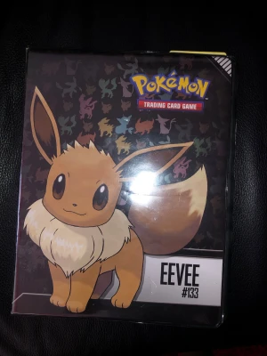 Pokémon Eevee samlarpärm Ultra Pro - Snygg Pokémon samlarpärm med Eevee-motiv och siluetter av olika Pokémon på svart bakgrund. Perfekt för att förvara och organisera trading cards. Officiell licens från Pokémon och Ultra Pro. Färgglada detaljer och robust plastmaterial. Pärmen har flera sidor till fullt av Pokémon kort av olika sorter 