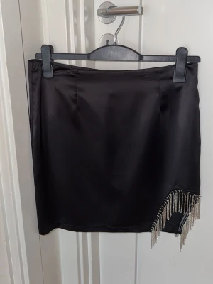 Svart kortkjol med pärlfransar - Snygg svart kortkjol i satinliknande material med glittriga pärlfransar på ena sidan. Kjolen är från Gina tricot och i storlek 40, men passar även 38💕