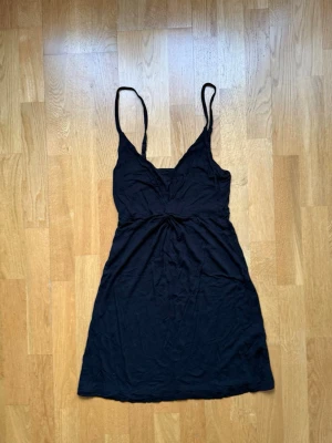 Svart kort klänning med v-ringning - Svart kort klänning med smala axelband och v-ringad hals. Klänningen har en twistad detalj i midjan som ger en snygg siluett. Perfekt för varma dagar eller när du vill ha en enkel men stilren look.