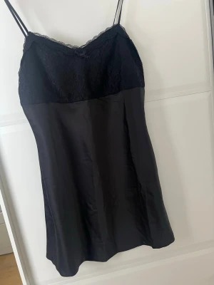 Svart nattlinne med spetsdetaljer - Snyggt svart nattlinne med tunna axelband och spets upptill. Överdelen har ett fint spetsmönster och en liten rosett vid urringningen, medan nederdelen är i slätt, glansigt tyg. Perfekt för en mysig kväll hemma.