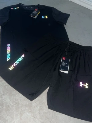 Svart träningsset från Under Armour - Snyggt svart träningsset från Under Armour med t-shirt och shorts. Båda plaggen har regnbågsfärgade loggor och textdetaljer. Perfekt för gymmet eller löprundan. Materialet är lätt och andas, vilket gör det bekvämt under träning.