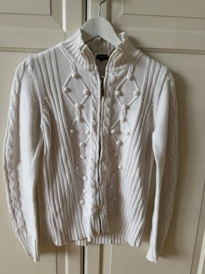 Vit stickad kofta från Lindex - Säljer en vit stickad kofta från Lindex med dragkedja framtill. Koftan har ett upphöjt mönster med bollar och ränder, samt ribbade muddar vid ärmslut och nederkant. 