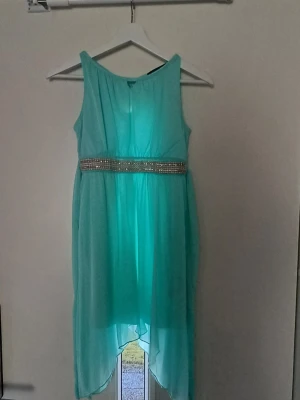 Turkos långklänning med pärlmidja - Snygg turkos långklänning med pärlbesatt midja och asymmetrisk nederkant. Klänningen är ärmlös och har knytning i ryggen för extra detalj. Perfekt för sommarens alla tillfällen.