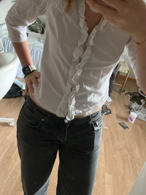 Vit blus med volang framtill - Säljer en vit blus med volangdetalj längs knäppningen framtill. Blusen har trekvartsärm och en feminin, figurnära passform. Perfekt för dig som gillar en klassisk men ändå trendig look. Perfekt för jobbet eller en lite mer uppklädd outfit.