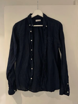 Mörkblå skjorta från Gant - Klassisk mörkblå skjorta från Gant med vita knappar och lång ärm. Skjortan har en enkel, stilren design och är tillverkad i ett mjukt material som känns bekvämt mot huden. Perfekt för dig som gillar tidlös stil.