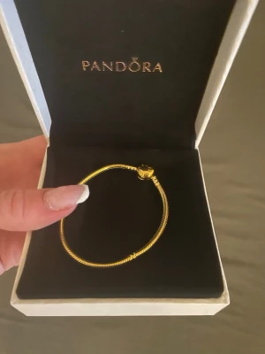 Pandora Armband - Snyggt guldpläterat armband från Pandora med klassiskt ormkedjemönster och ett elegant hjärtformat lås. Perfekt för att kombinera med dina favoritberlocker och skapa en personlig stil. Levereras i original ask.