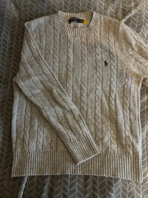 Grå kabelstickad tröja Polo Ralph Lauren - Säljer en grå kabelstickad tröja från Polo Ralph Lauren med rund halsringning och ribbade muddar. Tröjan har det klassiska broderade logotypen på bröstet och är tillverkad i mjukt garn. Perfekt för dig som gillar stilren och tidlös design. Inte använd mer än 2 gånger!