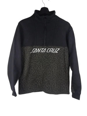 Santa Cruz Leopard Strip Quarter Zip Sweatshirt. S - Tröja från Santa Cruz med dragkedja halvvägs ner på framsidan. Tröjan har en svart överdel och en mönstrad underdel i mörkare toner. Santa Cruz-logotypen är placerad mitt på bröstet i vitt. Armhåla till armhåla: 53cm Axelbredd: 46cm Långt: 65cm Armlängden från armhåla: 45cm