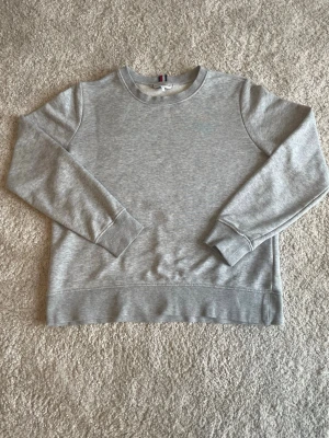 Grå sweatshirt från Tommy Hilfiger - Klassisk grå sweatshirt från Tommy Hilfiger med rund halsringning och långa ärmar. Tröjan har en liten broderad logga på ärmen och ribbade muddar vid ärmslut och nederkant. Perfekt basplagg för en avslappnad stil.
