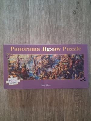 Panorama Jigsaw Puzzle 1000 bitar - Säljer ett panorama pussel med 1000 bitar i en lila kartong. Motivet visar en fantasifull stad i varma färger och storleken på pusslet är 98 x 33 cm. Perfekt för dig som gillar utmanande pussel och snygga motiv.