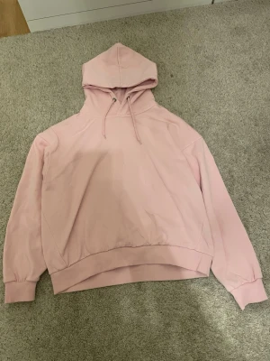 Ljusrosa hoodie med huva - Säljer en ljusrosa hoodie med huva och dragsnören. Hoodien har en avslappnad passform, långa ärmar och breda muddar vid ärmslut och nederkant. Perfekt för en chill och stilren look. Materialet är mjukt och skönt, troligtvis bomullsblandning.