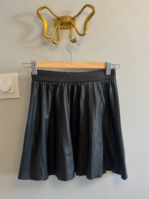 Svart kortkjol i skinnimitation - Säljer en svart kortkjol med lätt utställd passform och bred midjeresår. Kjolen har en snygg skinnlook