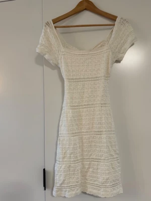 Vit spetsklänning med öppen rygg - Söt vit kortklänning i spets med korta ärmar och öppen rygg. Klänningen har ett fint spetsmönster över hela och en snygg twistdetalj bak. Perfekt för sommarens alla tillfällen!