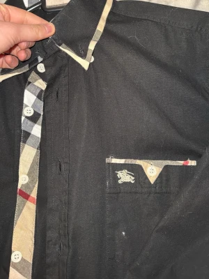 Svart skjorta med rutiga detaljer Burberry - Snygg svart skjorta från Burberry med klassiska beige, röda och svarta rutiga detaljer på krage,  manschetter och bröstficka. Skjortan har vita knappar och en broderad logga på fickan. Perfekt för dig som gillar stilrena plagg med en twist. Storlek S