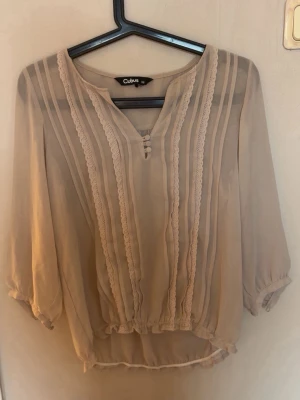 Beige blus med spets från Cubus - Söt beige blus från Cubus med trekvartsärm och v-ringning. Blusen har fina spetsdetaljer och knappar framtill samt lätt transparent material. Perfekt att styla med jeans eller kjol för en trendig look.