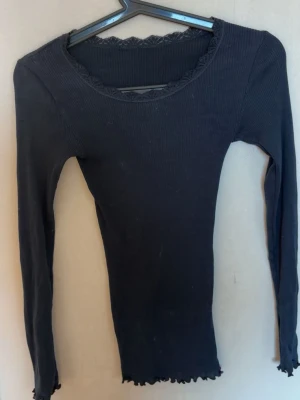 Svart ribbad långärmad topp - Säljer en svart ribbad långärmad topp med spetsdetalj vid halsringningen och vågig kant nertill. Perfekt att styla med jeans eller kjol för en trendig look. Superenkel att matcha och passar till många olika outfits.