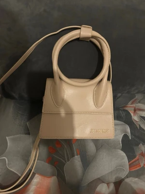 Beige Jacquemus Le Chiquito väska - Säljer en beige Jacquemus Le Chiquito handväska i slätt läder med rundade handtag och axelrem. Väskan har en stilren, boxig form och diskret Jacquemus-logga i guld på framsidan. Perfekt för dig som gillar trendiga accessoarer.